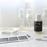  Prolong Lash cleanser products arranged on a white tiled surface with foaming bottles, a concentrate bottle, instruction cards, and cleansing brushes. 