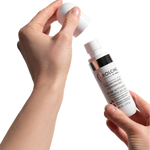 A close-up of a person's hands holding a white bottle of PROLONG Lash Eyelash Hygiene Cleansing Mist and lifting its cap against a solid white background.